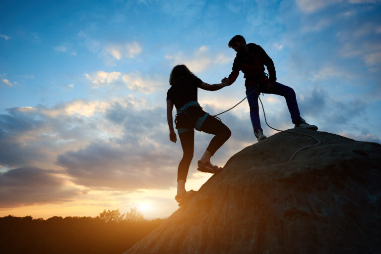 Two people rock climbing outdoors at sunset, with one climber helping the other up a rock face.